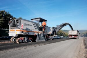Wirtgen cold milling machine removing asphalt during a road rehabilitation project while loading material into a dump truck.